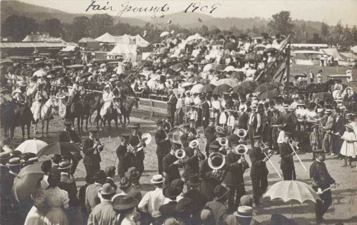 Black and white photograph of the Ulster County Fair parade in 1909, showing the midway and Ferris wheel in the background. Marching in the parade is Clayton’s Military Band, followed by a row of horseback riders. Spectators are standing, walking, and seated in the grandstand.